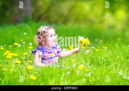 Entzückende Kleinkind Mädchen mit dem lockigen Haar trägt ein rosa Kleid mit gelben Löwenzahn in einen blühenden Frühling Garten Stockfoto