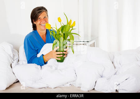 Frau in den Fünfzigern im Bett sitzen und halten eine Vase mit Blumen Stockfoto