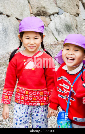 Zwei japanische Schüler, Junge und Mädchen, 6-7 Jahre alt, beide tragen rote Schuloberteile mit violetten Kappen, betrachten Zuschauer, Mädchen lächeln, Junge lachen. Stockfoto