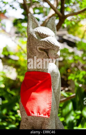 Goscha-Inari-Schrein in Kobe, Japan. Nahaufnahme einer steinernen Kitsune-Statue, eines Fuchskopfes, der einen Ball im Mund hält, und eines roten Lätzchens um den Hals. Stockfoto