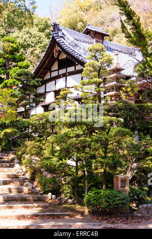 Goscha-Inari-Schrein in Kobe, Japan. Breite Steinstufen, die zur Haupthalle, Hondo, führen, auf einem kleinen Hügel mit Steinpagode unter den Bäumen. Helles Sonnenlicht. Stockfoto