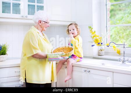 Glücklich schöne Urgroßmutter und liebenswert Enkelin, geschweiften Kleinkind Mädchen im bunten Kleid, einen Apfelkuchen backen Stockfoto