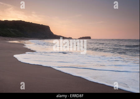 Ruhige See, plätschernden Wellen, weichen Sand, Ufer, Silhouette Klippen & gegen Nab unter roten Himmel bei Sonnenuntergang - malerischen ruhigen gegen Bucht, Yorkshire, Großbritannien. Stockfoto