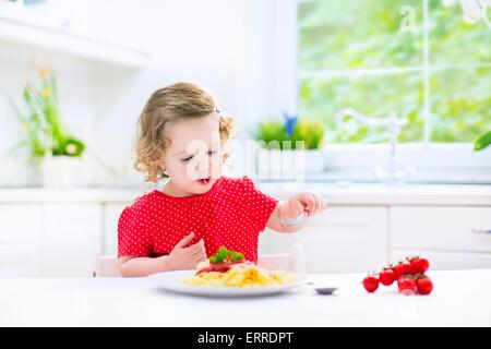 Niedliche lockige lachende Kleinkind Mädchen in ein rotes Hemd mit Gabel und Löffel essen Spaghetti mit Tomatensauce und Gemüse spielen Stockfoto