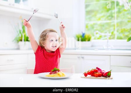 Niedliche lockige lachende Kleinkind Mädchen in ein rotes Hemd mit Gabel und Löffel essen Spaghetti mit Tomatensauce und Gemüse spielen Stockfoto