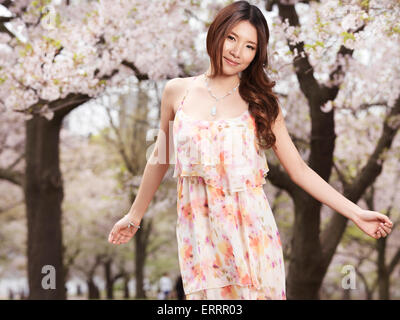 Junge lächelnde glücklich Asiatin in einem geblümten Kleid in einem Park mit blühenden Chery Bäume im Frühling Stockfoto