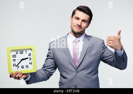 Geschäftsmann hält Uhr und zeigt Daumen über grauen Hintergrund Stockfoto