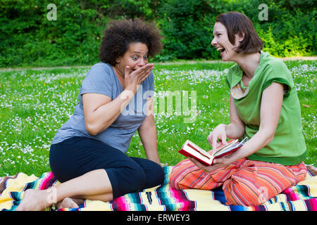 zwei Frauen sitzen auf Decke im Park mit einem Buch und lachen Stockfoto