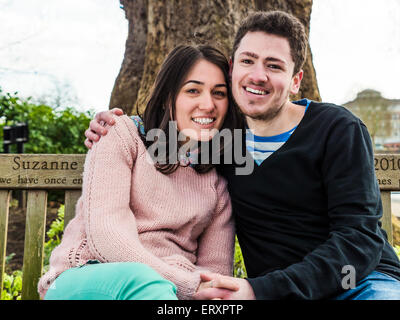 Romantische junges Paar auf Parkbank beisammen sitzen und Lächeln in die Kamera schaut Stockfoto