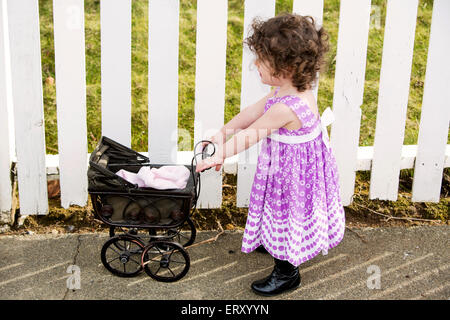 kleines Mädchen schob einen Vintage Kinderwagen vor einem weißen Zaun Stockfoto