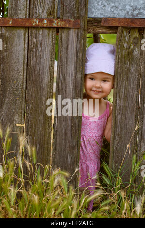 Kleine Mädchen spielen Peek ein Boo durch eine Lücke in einer gebrochenen Plank in einem rustikalen hölzernen Zaun Stockfoto