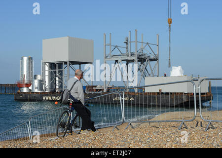 Ein Mann saß auf einem Fahrrad am Strand beobachtet, wie ein Kran Teil der British Airways ich 360 Turm entlädt aus ein Schiff im Meer während der aussichtstürme Bau in Brighton, Sussex, UK.