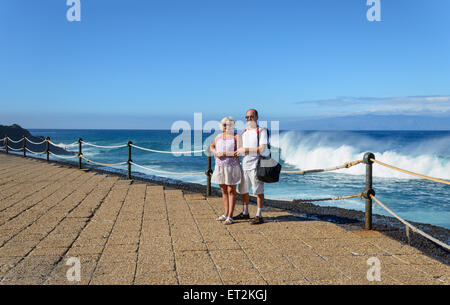 Älteres gegerbt paar in leichter Kleidung steht am Ufer in der Sonne am strahlend blauen Wasser und Himmel und Spritzwasser winken zurück Stockfoto