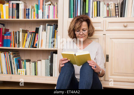 Ältere Frau sitzt auf dem Boden vor Bücherregal und Lesung, München, Bayern, Deutschland Stockfoto