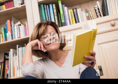 Ältere Frau sitzt vor Bücherregal und Lesung, München, Bayern, Deutschland Stockfoto