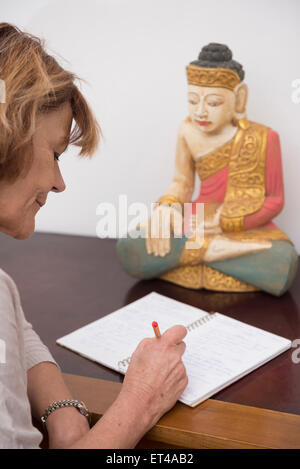 Ältere Frau schreiben in Notebook, Buddha-Statue im Hintergrund, München, Bayern, Deutschland Stockfoto
