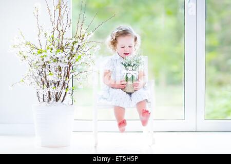 Schöne lockige Kleinkind Mädchen in einem weißen Kleid sitzt in einem weißen Schaukelstuhl mit ersten Frühlingsblumen in Kristallvase Stockfoto