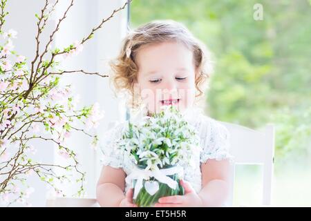 Schöne lockige Kleinkind Mädchen in einem weißen Kleid sitzt in einem weißen Schaukelstuhl mit ersten Frühlingsblumen in Kristallvase Stockfoto