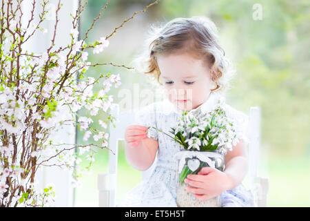 Schöne lockige Kleinkind Mädchen in einem weißen Kleid sitzt in einem weißen Schaukelstuhl mit ersten Frühlingsblumen in Kristallvase Stockfoto