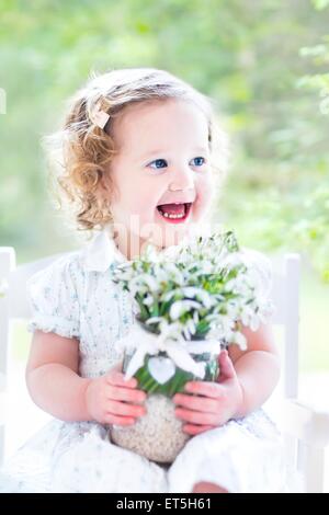 Schöne lockige Kleinkind Mädchen in einem weißen Kleid sitzt in einem weißen Schaukelstuhl mit ersten Frühlingsblumen in Kristallvase Stockfoto