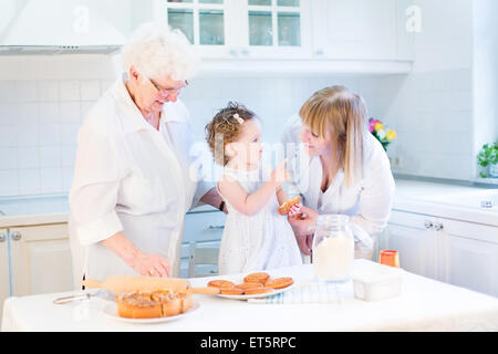 Niedliche lockige Kleinkind Mädchen Backen einen Kuchen mit ihrer Großmütter in eine weiße schöne Küche Stockfoto