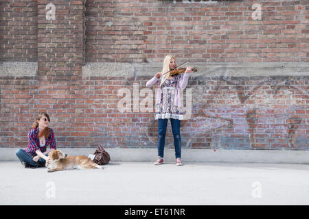 Frau sitzt mit ihrer Freundin spielt Geige vor Mauer, München, Bayern, Deutschland Stockfoto