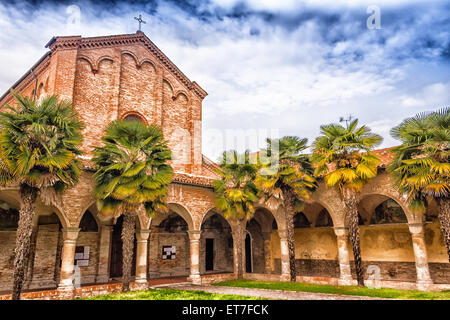 Brickwall Fassade und Kolonnade mit Bögen der XV Jahrhundert römische gotische Kirche Saint Francis in Cotignola in der Nähe von Ravenna in der Landschaft der Emilia Romagna in Italien gewidmet. Reihen von Palmen säumen den Garten. Stockfoto