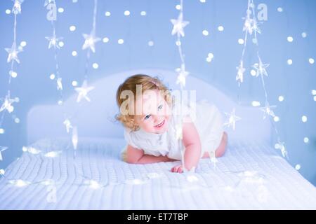 Entzückende Kleinkind Mädchen mit dem lockigen Haar tragen ein wunderschönes weißes Kleid spielen in einem Schlafzimmer mit Weihnachtsbeleuchtung Stockfoto