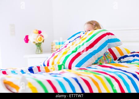 Lustiges kleines Kleinkind Mädchen sitzt in einem weißen Bett mit regenbogenfarbenen Streifen Kissen und decken Blatt spielen verstecken und suchen Stockfoto