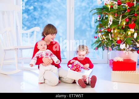 Drei Kinder öffnen Weihnachtsgeschenke, Weihnachten Morgen in ein Wohnzimmer mit geschmückten Baum und Fenster in einem verschneiten Garten genießen Stockfoto