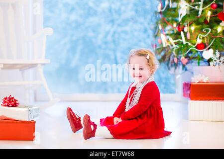 Niedliche lockige kleine Mädchen in einem roten Kleid und weiße Perlenkette spielen unter einem Weihnachtsbaum mit präsentiert auf dem Boden sitzend Stockfoto