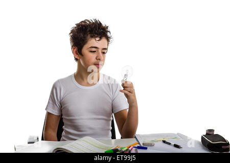 Junge ist Hausaufgaben: sitzen vor einem Schreibtisch von Büchern, Heften und Stiften abgedeckt, er eine Glühbirne mit seiner linken Hand hält und mit der Befragung Augen Uhren Stockfoto