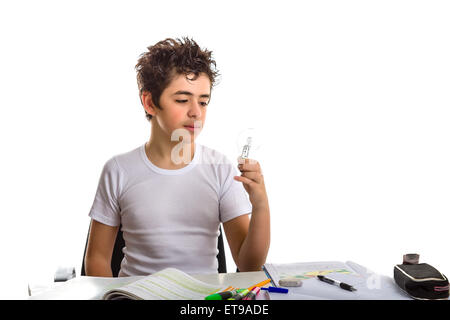 Junge ist Hausaufgaben: sitzen vor einem Schreibtisch von Büchern, Heften und Stiften abgedeckt, er eine Glühbirne mit seiner linken Hand hält und mit der Befragung Augen Uhren Stockfoto