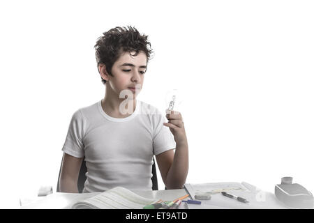 Junge ist Hausaufgaben: sitzen vor einem Schreibtisch von Büchern, Heften und Stiften abgedeckt, er eine Glühbirne mit seiner linken Hand hält und mit der Befragung Augen Uhren Stockfoto