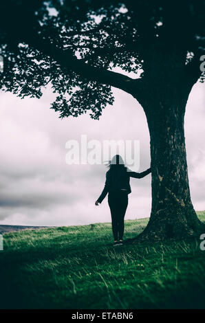Silhouette einer weiblichen Figur lehnt sich an einen Baum in der Landschaft Silhouette einer weiblichen Figur lehnt sich an einen Baum in der Landschaft