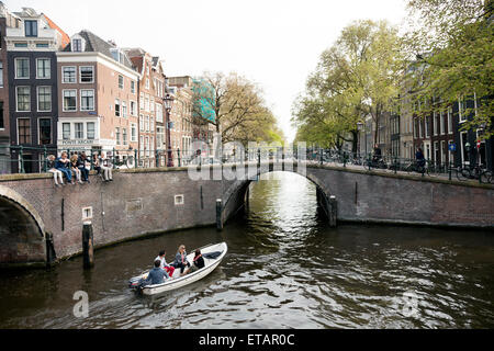 junge Leute sitzen auf Brücke über Amsterdam Canal in der Nähe von Rembrandt Plein im Frühjahr während kleines Boot vorbeifährt Stockfoto