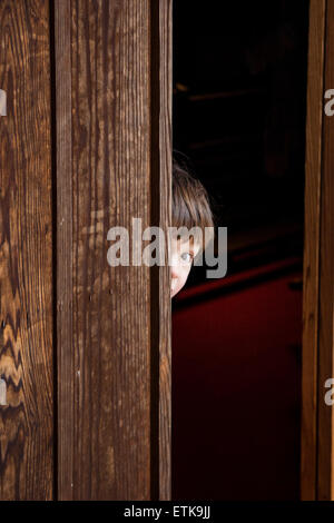 Kind, Junge, 4-5 Jahre alt, hinter der alten Holztür herausprießend. Die Hälfte des Gesichts und des Kopfes ist versteckt, ein Auge macht Blickkontakt mit dem Betrachter. Stockfoto