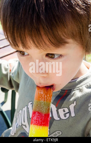 Oben nah am Gesicht des Kindes, Junge, 4-5 Jahre alt, beißt sich in einen mehrfarbigen Eislastwagen, während es im Freien bei Sonnenlicht liegt. Stockfoto