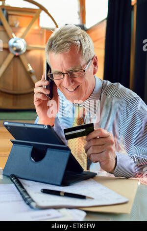 Online Kreditkarte reife Kaufmann in seinem Hausboot Hausboot Büro damit eine Karte Transaktion auf Linie mit dem iPad Tablet Computer & iPhone Smartphone Stockfoto