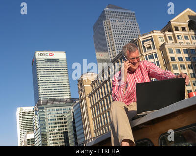 Reifer Mann auf seinem Boot am iPhone Smartphone Blick auf seinem Laptop Computer Canary Wharf Büros hinter London E14 sprechen außerhalb arbeiten Stockfoto