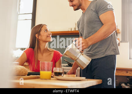 Aufnahme des jungen Paares frühstücken in Küche. Junger Mann stehen und servieren von Kaffee mit Frau sitzen am Frühstückstisch Stockfoto