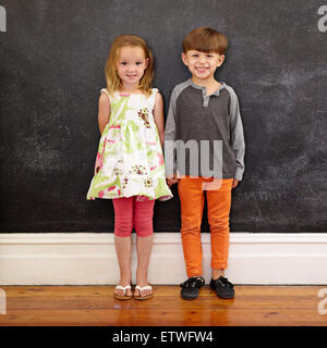 Voller Länge Schuss von zwei kleinen Kindern zusammenstehen gegen eine Tafel. Kleinen Jungen und Mädchen zu Hause Blick in die Kamera Stockfoto