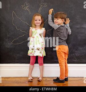 In voller Länge Portrait von niedliche kleine Mädchen stehen und kleiner Junge Engelsflügel um sie herum auf Tafel zeichnen. Zwei kleine Kinder s Stockfoto