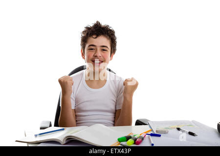 Kaukasischer Junge sitzt vor Hausaufgaben: er freut sich und macht eine Geste des Erfolgs Hände erhebend Stockfoto