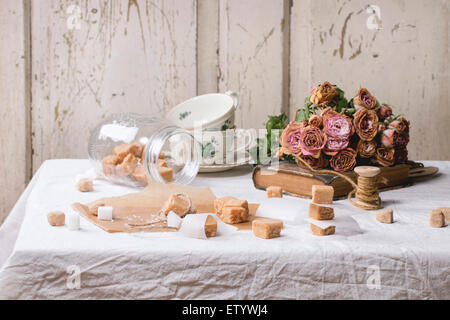 Fudge, Süßigkeiten und Karamell auf Backpapier und in Glas, diente über weiße Tischdecke mit Bouquet von trockenen rosa Rosen und thread Stockfoto