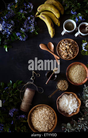 Zutaten für Müsli Banane Brot sind auf einer dunklen Oberfläche angezeigt, umgeben von frischen Frühlingsblumen. Stockfoto