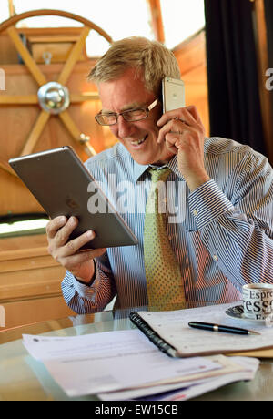 IPad iPhone Online-Meeting Geschäftsmann im Barge Houseboat Büro mit Blick auf seinen iPad Air Tablet Smart Computer Bildschirm und mit seinem iPhone Smartphone Stockfoto