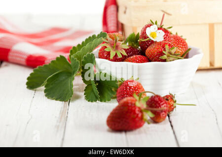 reife Erdbeeren in und vor kleinen Schüssel verziert mit Blättern und Blüten, Korb und Küche Handtuch im Hintergrund Stockfoto