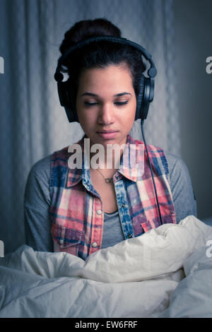 Eine junge Frau hört Kopfhörer in ihrem Zimmer unter der Bettdecke. Stockfoto