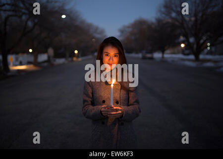 Eine asiatische Mädchen in einem Mantel hält eine Kerze in der Abenddämmerung in der Straße von einer Kleinstadt-Umgebung. Stockfoto